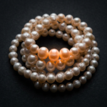 A coiled strand of pearls varying in size and sheen, glowing with warm orange light where the strand curves, resting against a dark background.